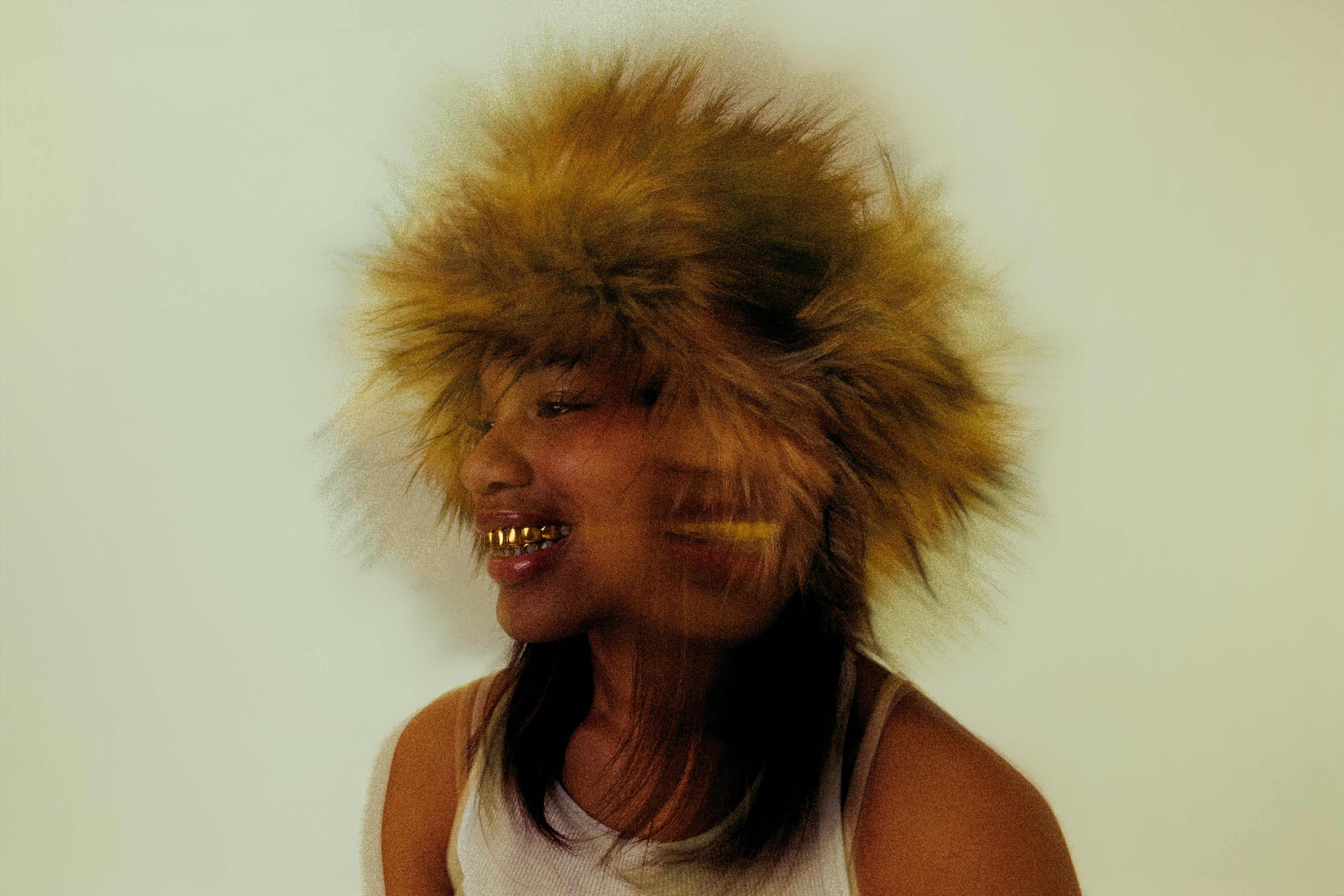 Profile shot of a woman smiling, wearing a fur hat and gold teeth grills against a light backdrop, creating a unique fashion statement.
