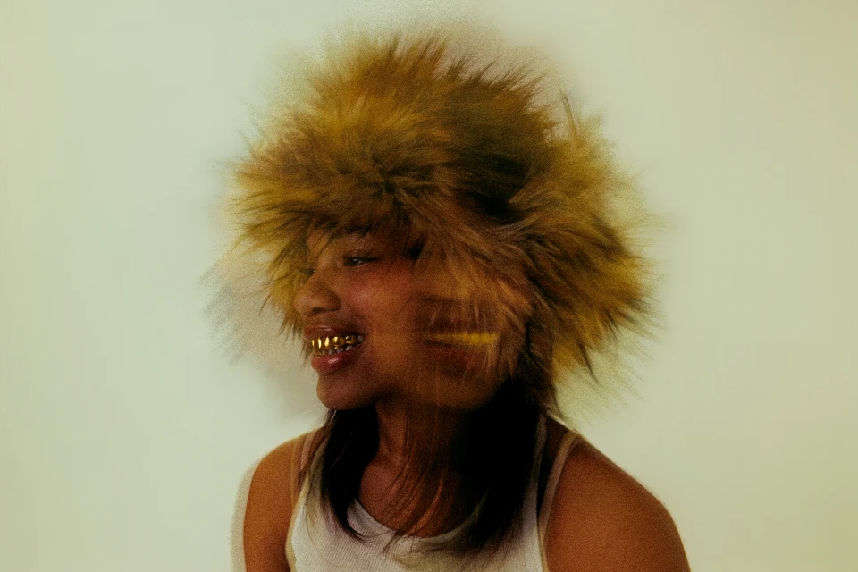 Profile shot of a woman smiling, wearing a fur hat and gold teeth grills against a light backdrop, creating a unique fashion statement.