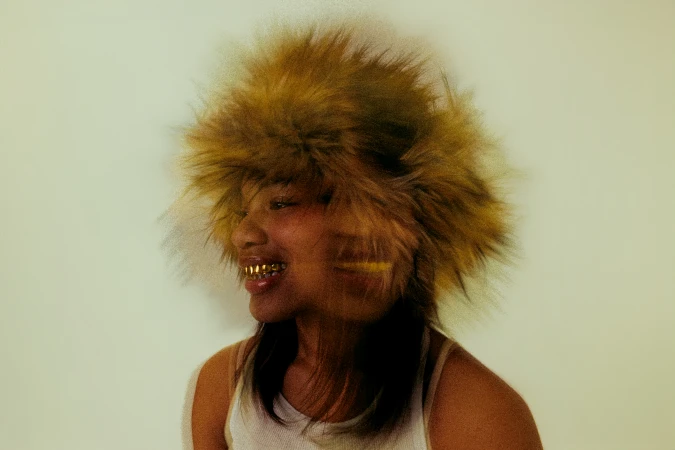 Profile shot of a woman smiling, wearing a fur hat and gold teeth grills against a light backdrop, creating a unique fashion statement.