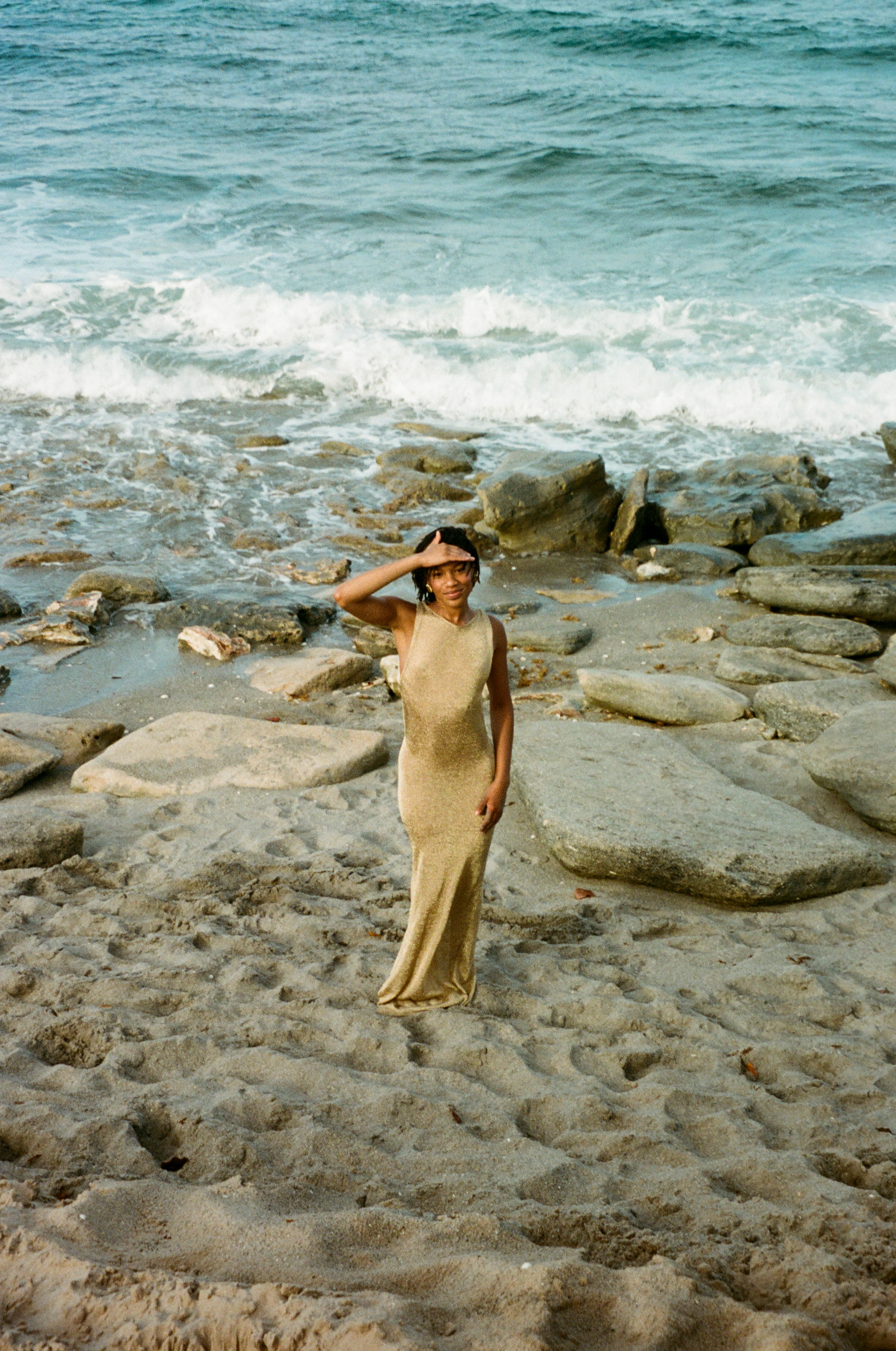 A woman in a dress stands on a beach with rocks, shielding her eyes from the sun, with the ocean and waves in the background.