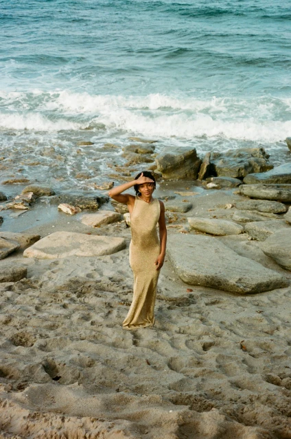 A woman in a dress stands on a beach with rocks, shielding her eyes from the sun, with the ocean and waves in the background.