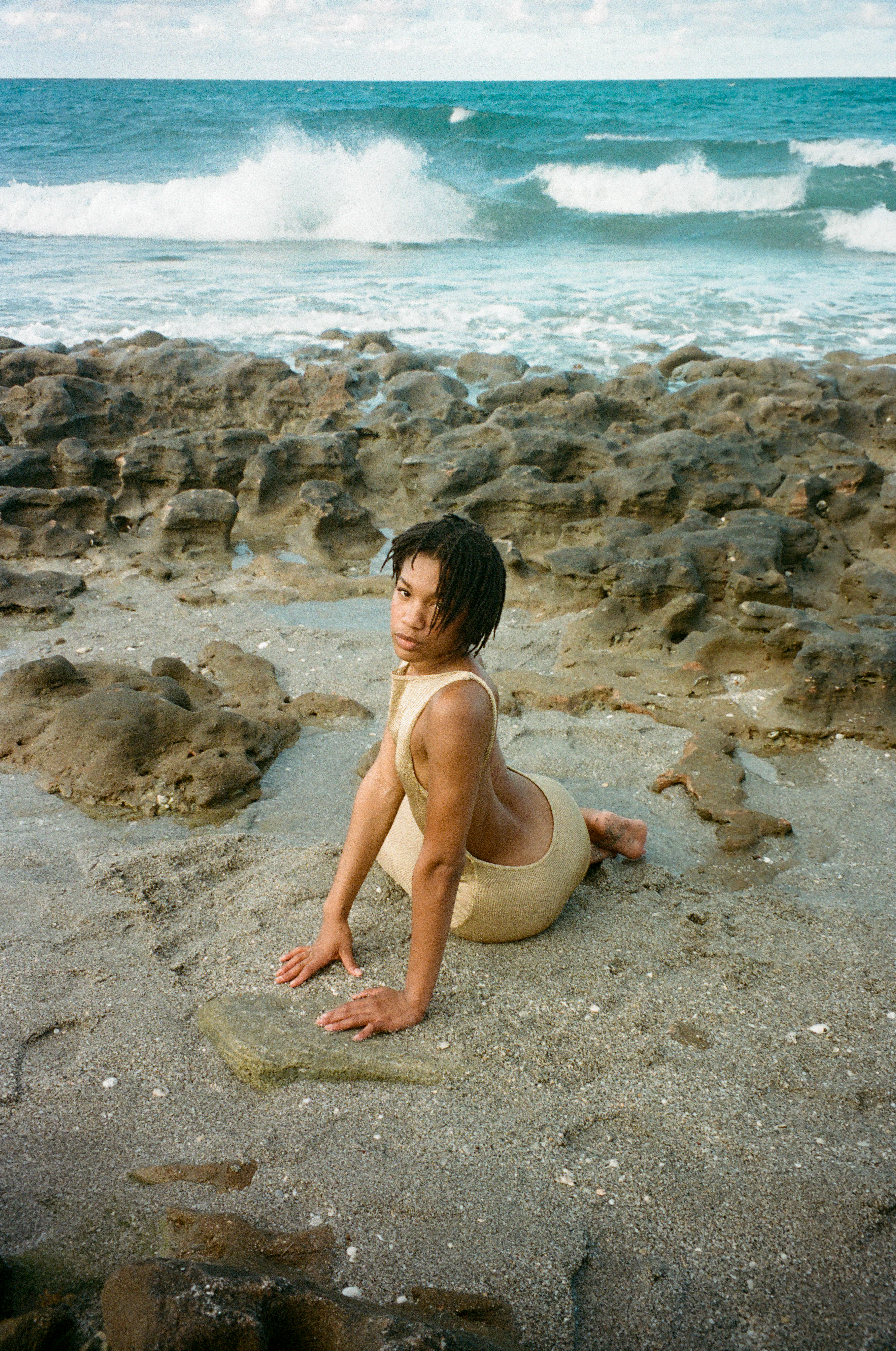 Woman in swimwear poses on rocky beach with ocean waves. Low angle shot captures tan one-piece swimsuit and weathered rock texture.