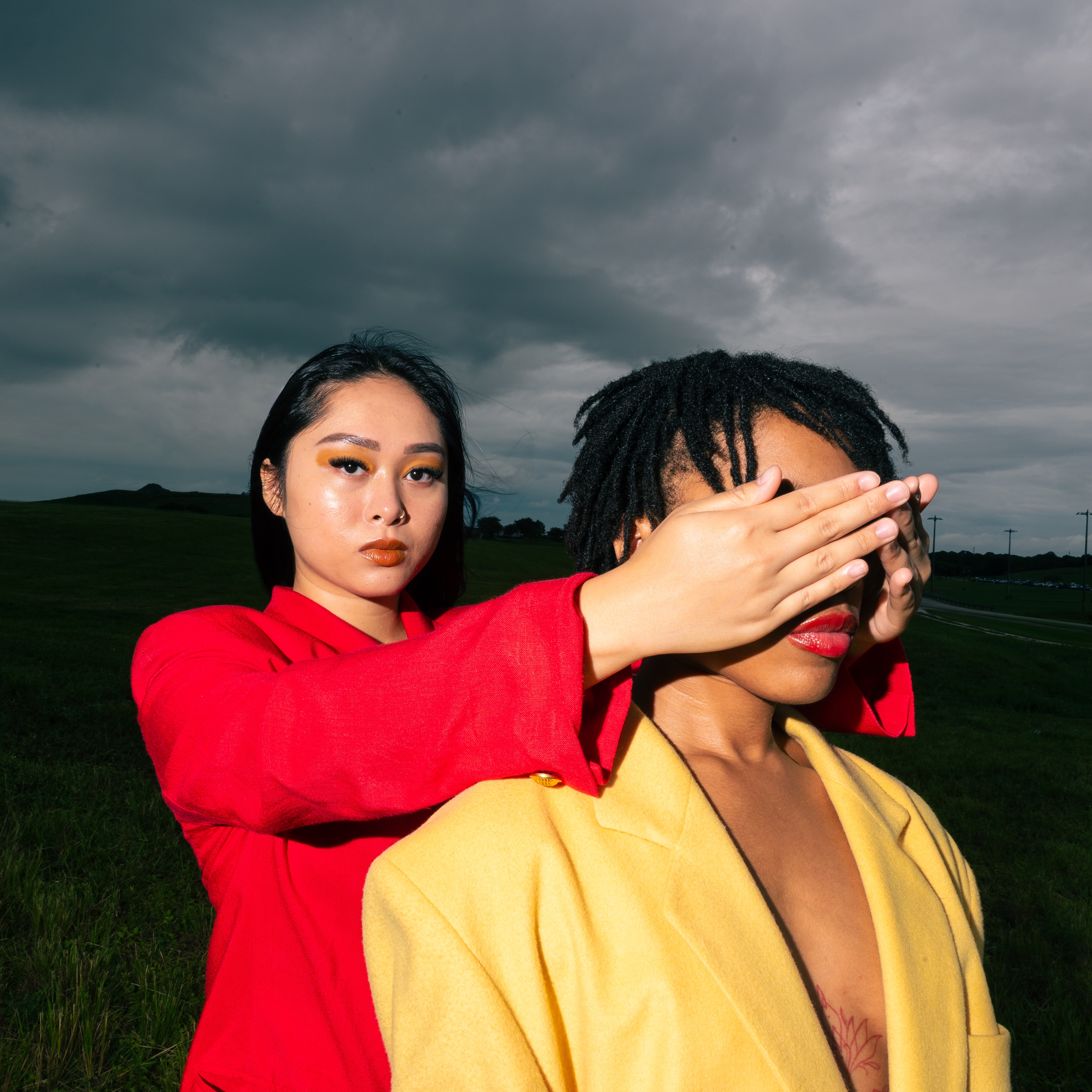 Two people in bright jackets pose in a field under a cloudy sky. One person covers the eyes of the other with their hands.