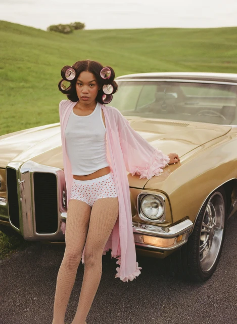 Young woman with curlers poses by a gold vintage car on a green hill. She wears a pink robe, white top, and polka-dot shorts.