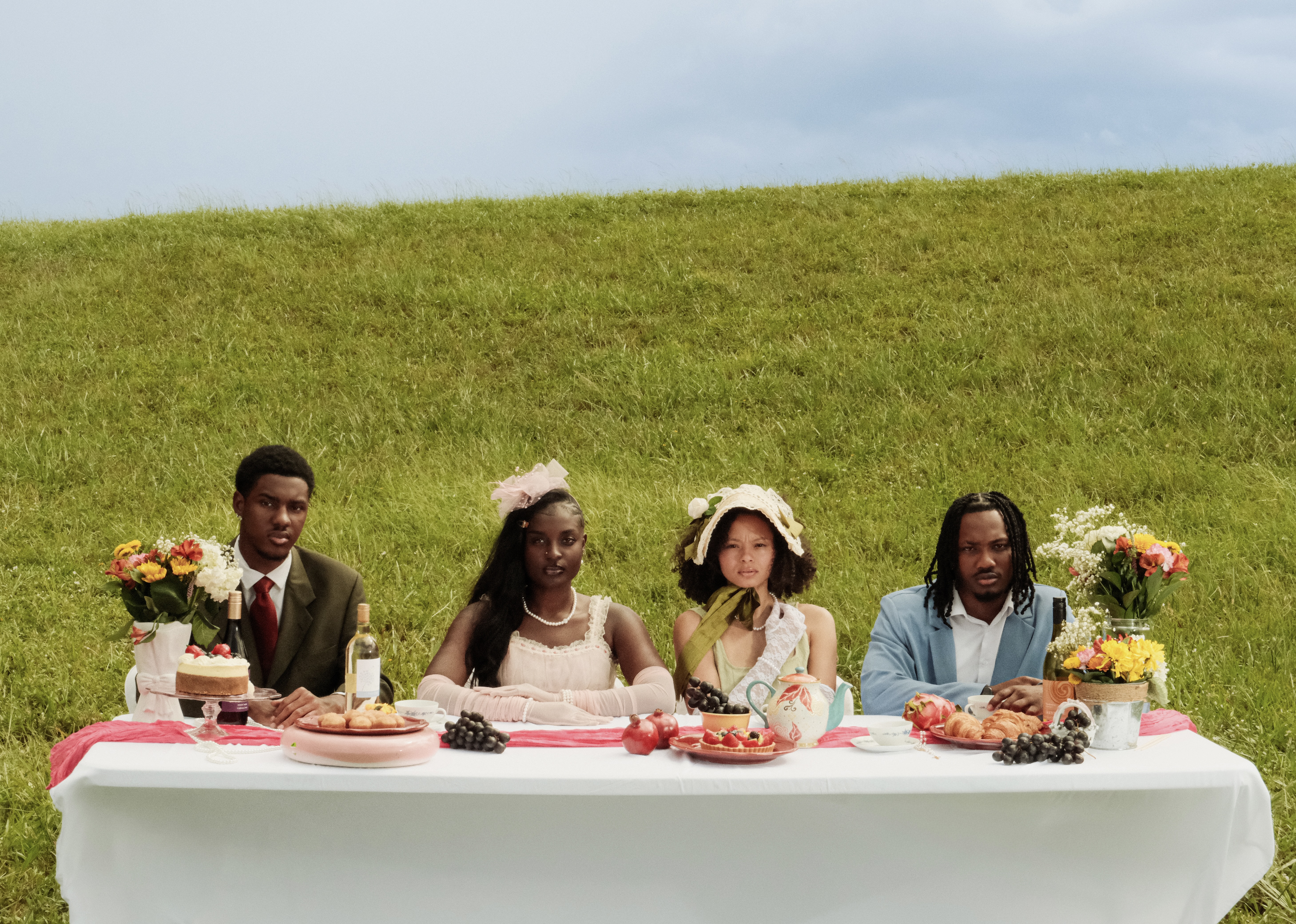 Four people seated at a table in a grassy field. The table is set with food and flowers, and the sky is visible in the background.