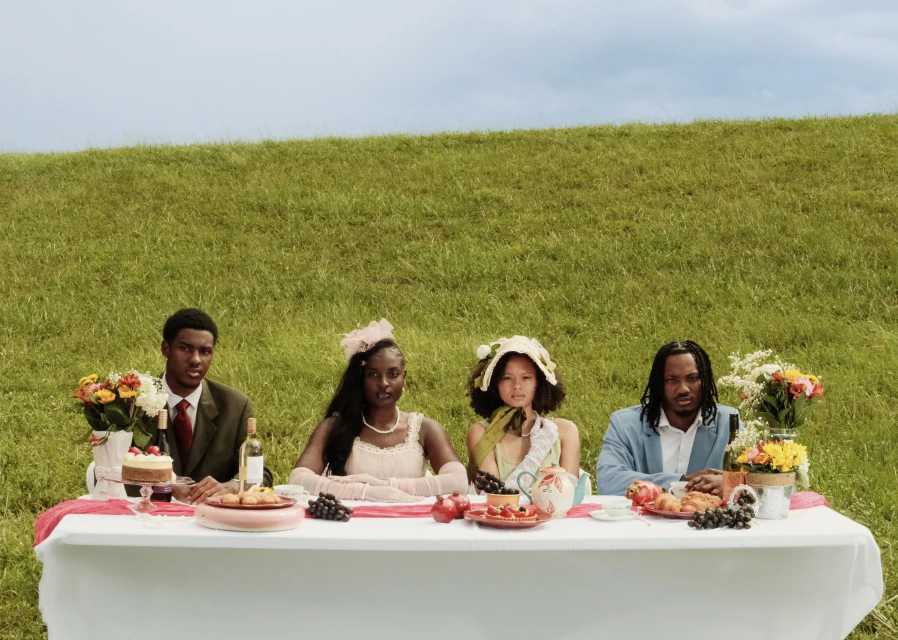 Four people seated at a table in a grassy field. The table is set with food and flowers, and the sky is visible in the background.