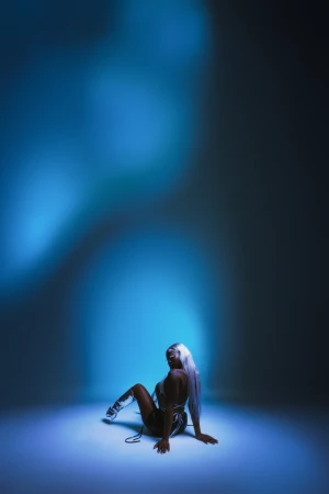 A woman with long white hair poses on the floor in metallic apparel, looking over her shoulder in a studio with cool blue lighting.
