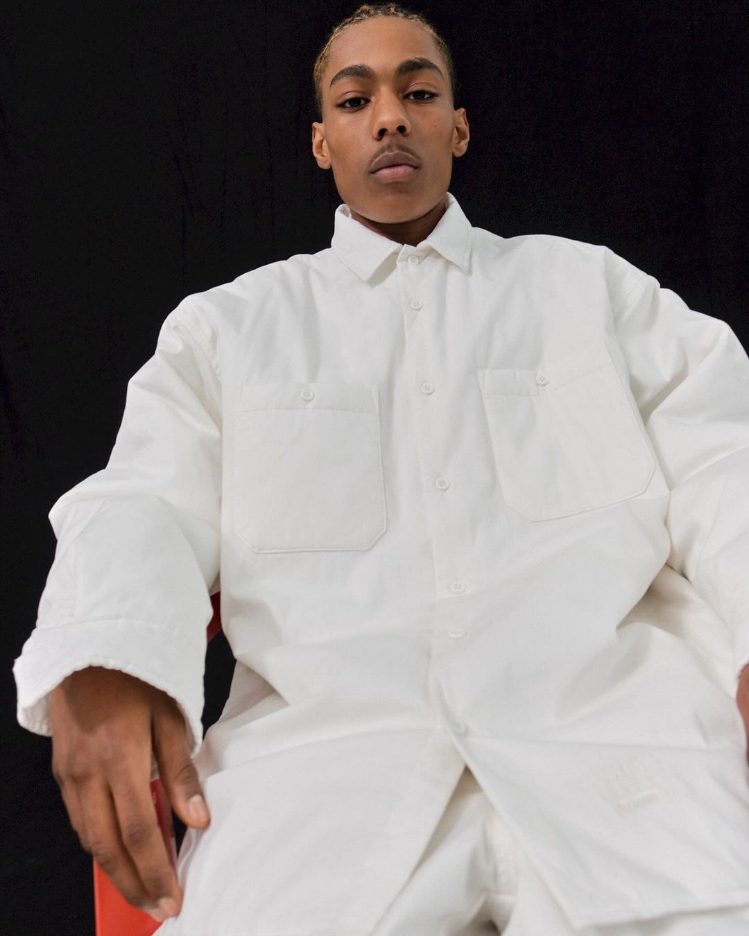 Portrait of an adult Black male in a white button-down shirt seated on a red chair against a black background, shot at eye-level.