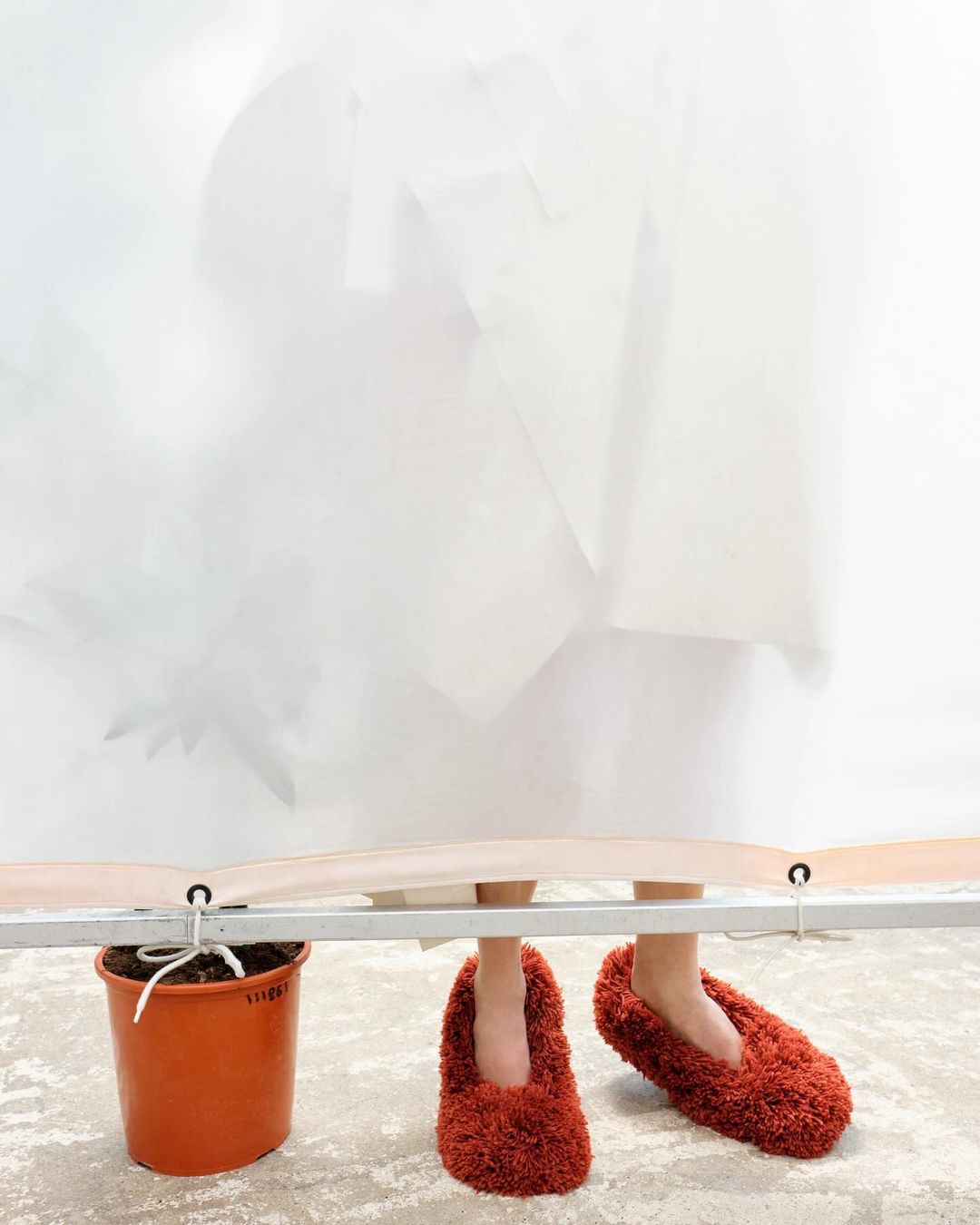 A person standing behind a white screen wearing oversized fuzzy red slippers next to a potted plant in a bright indoor setting.
