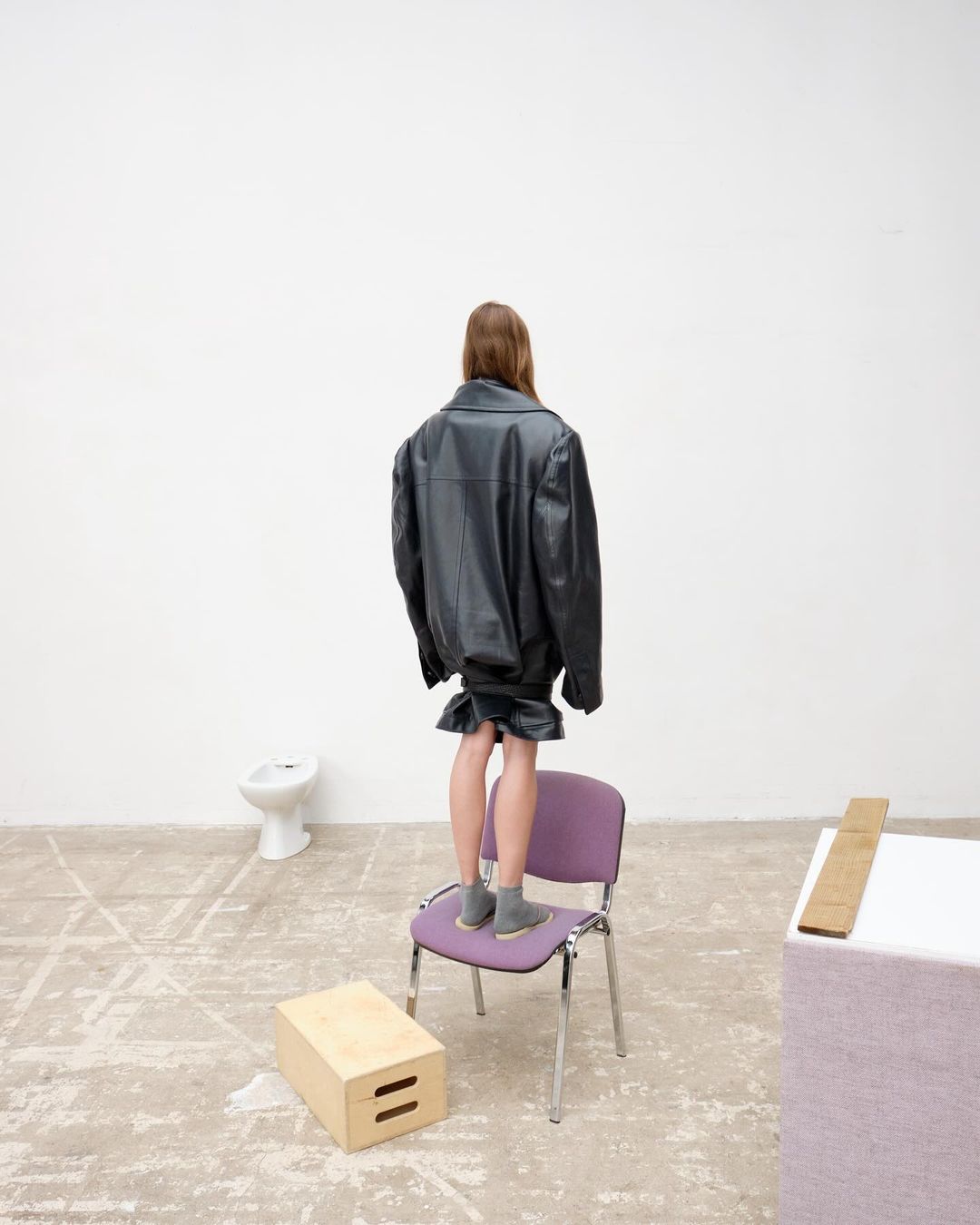 A person stands on a chair in an oversized black leather jacket and matching skirt in an empty room with a toilet in the background.