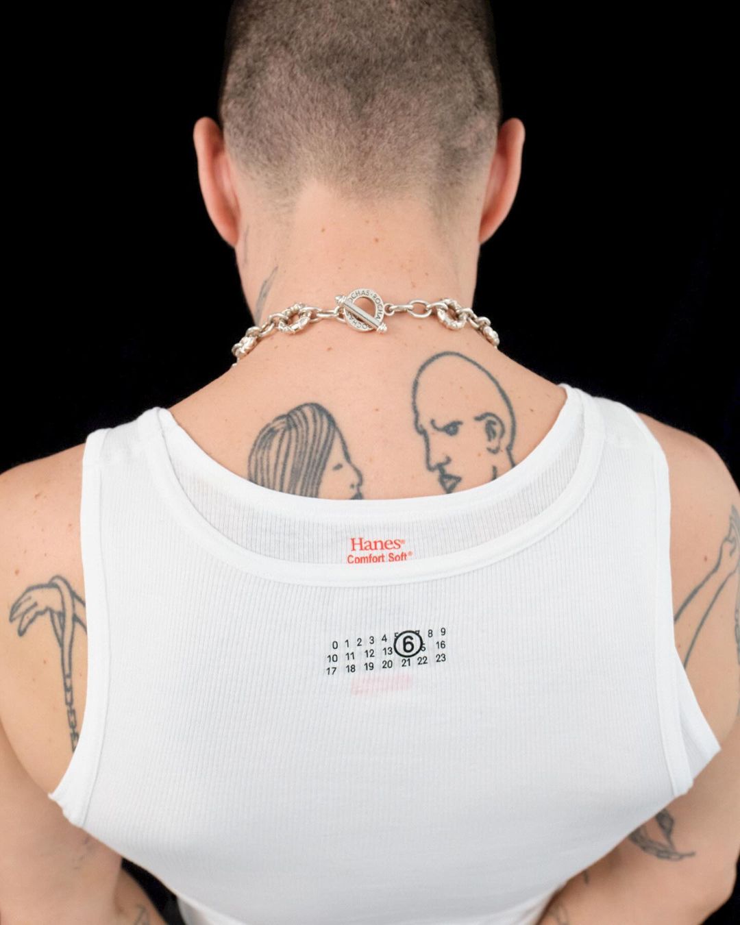 Man shows back wearing white tank top, tattoos, and chunky silver necklace against black backdrop in studio lighting.