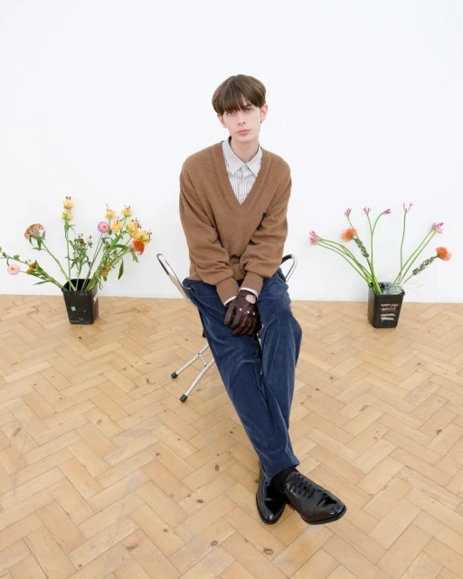 Young man in V-neck sweater poses on metal chair between two flower arrangements in indoor studio with light hardwood floor.