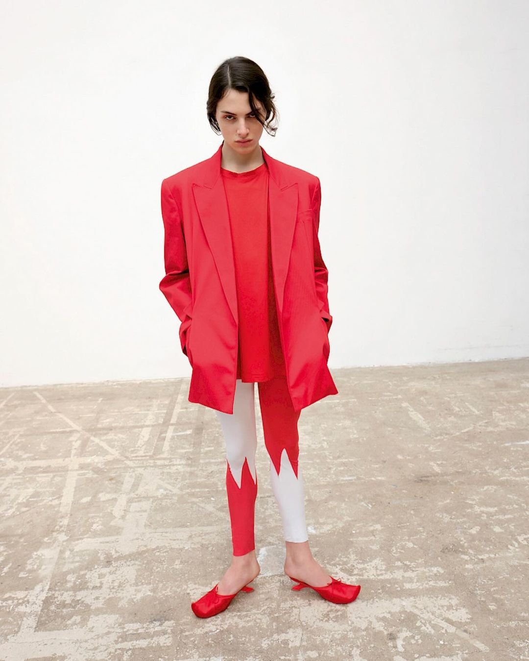 Fashion shot of a young adult woman in a red blazer, T-shirt, and leggings with red heels, posing against a neutral studio background.