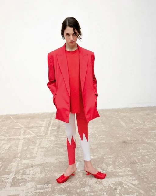 Fashion shot of a young adult woman in a red blazer, T-shirt, and leggings with red heels, posing against a neutral studio background.