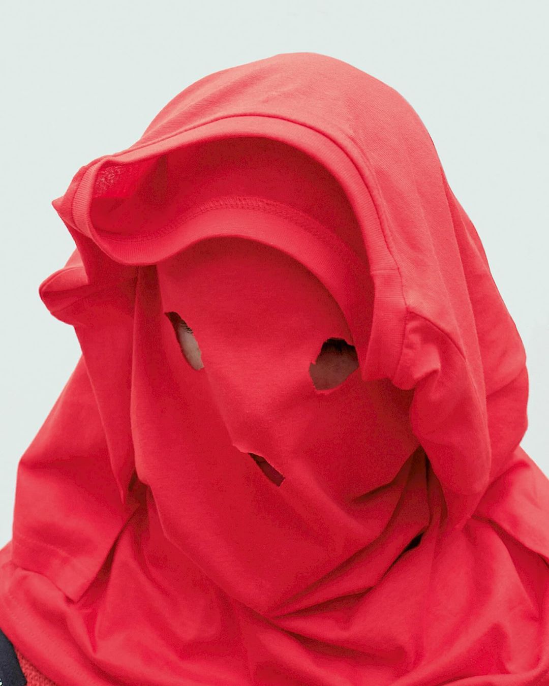 Person wearing a red t-shirt over their head with holes cut out for the eyes and mouth, posing against a white background.