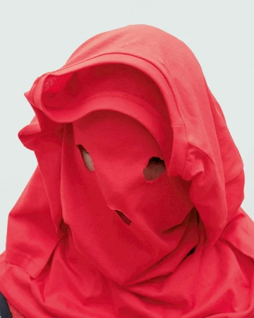 Person wearing a red t-shirt over their head with holes cut out for the eyes and mouth, posing against a white background.