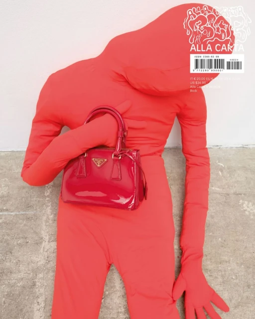 Person in red jumpsuit holds a red Prada purse against a white wall, presented as an editorial fashion statement.