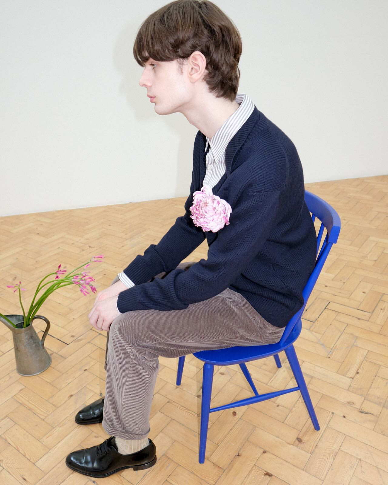 Young man in casual attire sits on blue chair with flower accent, indoors on herringbone parquet floor. Fashion forward, calm demeanor.