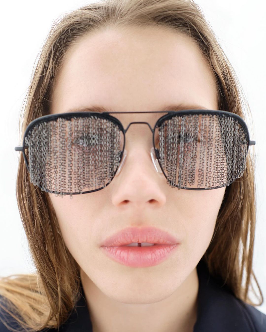 Close-up portrait of a young woman wearing artistic sunglasses with a mesh pattern in a minimalist indoor setting with bright, neutral lighting.