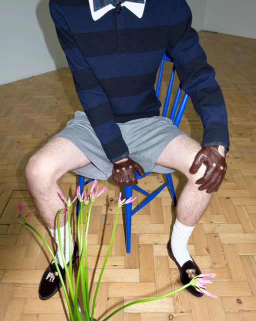 A person sits on a blue chair wearing a striped rugby shirt, gray shorts, brown leather gloves, dress socks, and loafers, with flowers in front.
