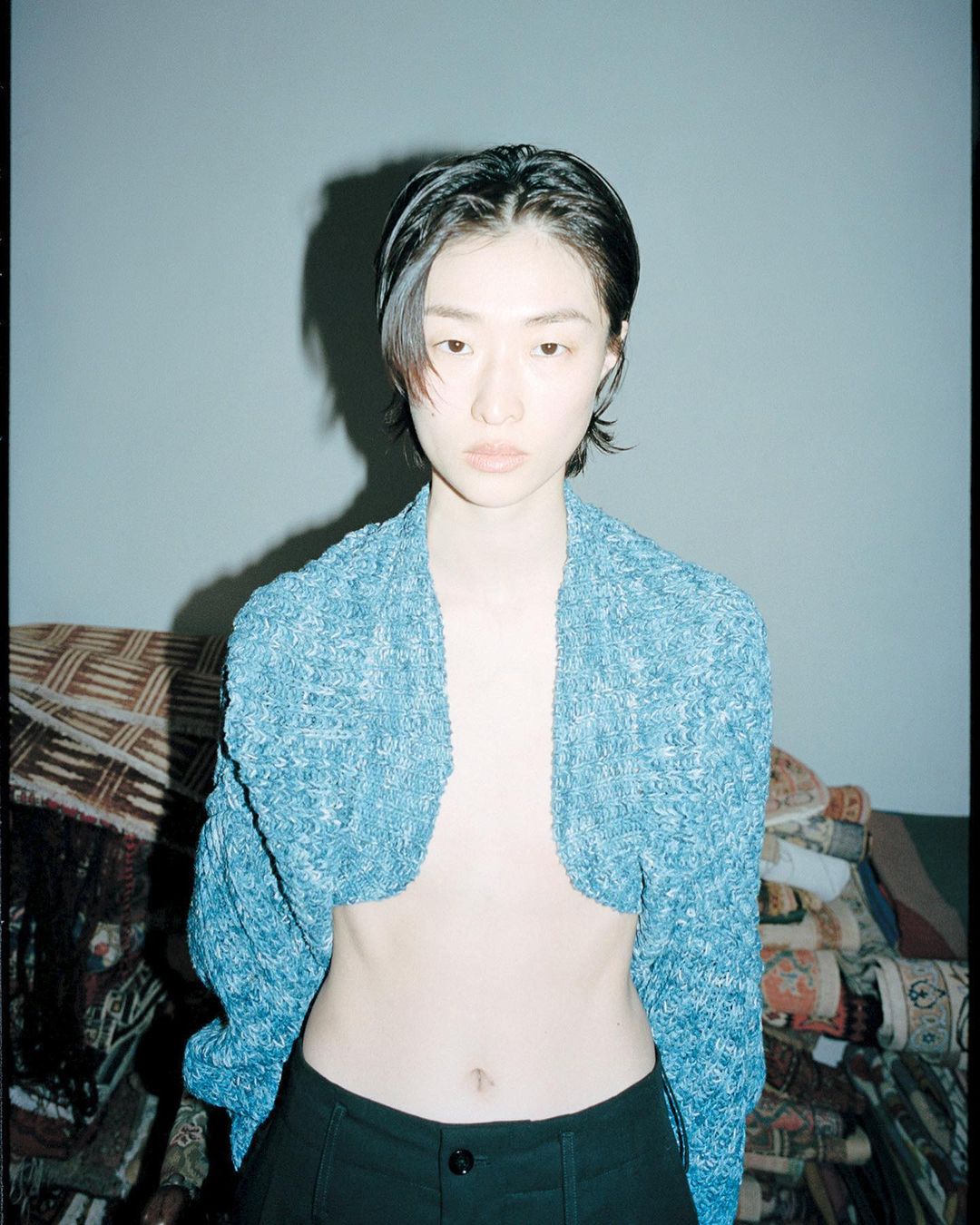 Close-up portrait of a young adult woman with short black hair wearing a blue knitted cardigan and black pants in an indoor setting.