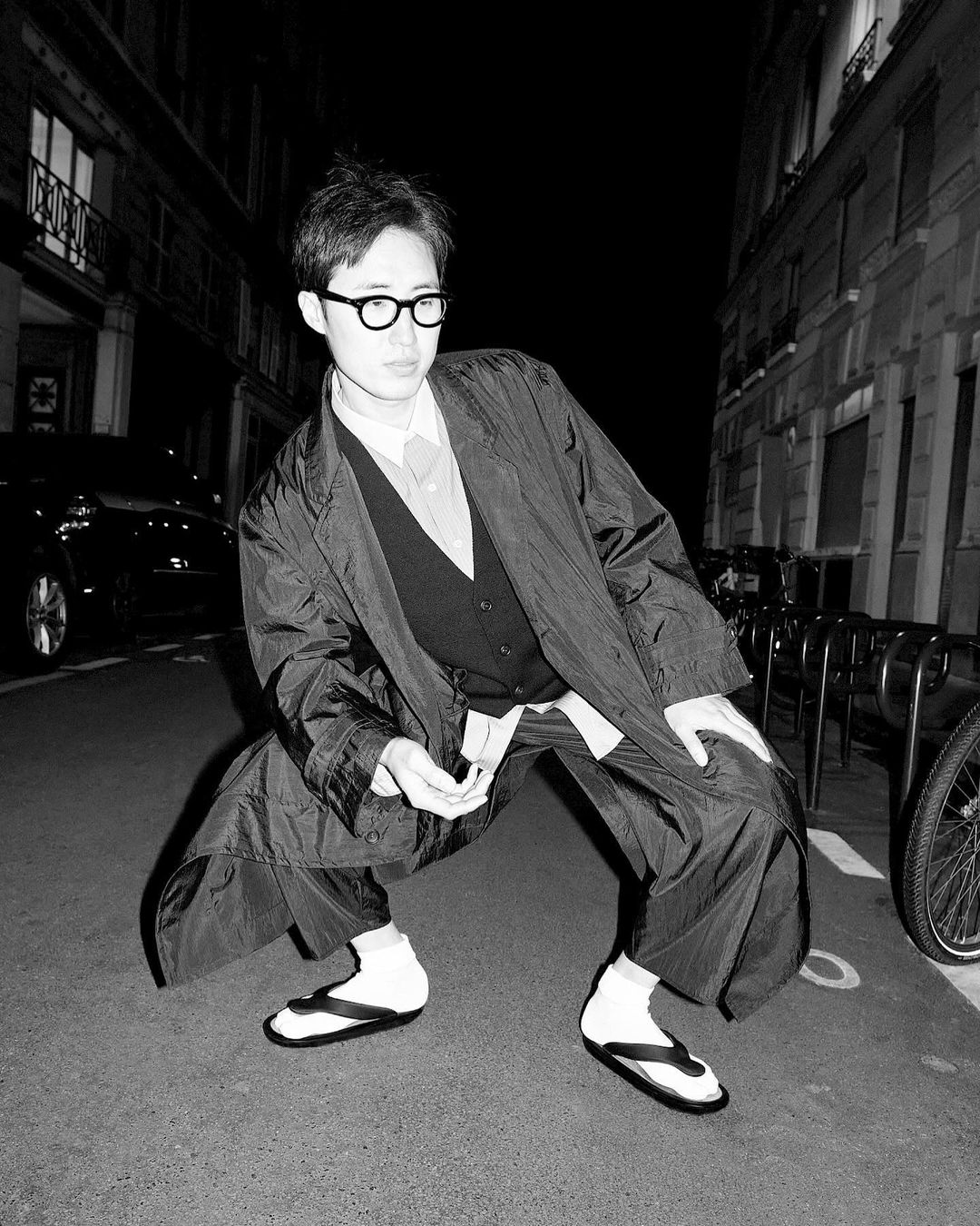 Black-and-white street style portrait of a young adult wearing glasses and a dark jacket crouching in an urban environment.
