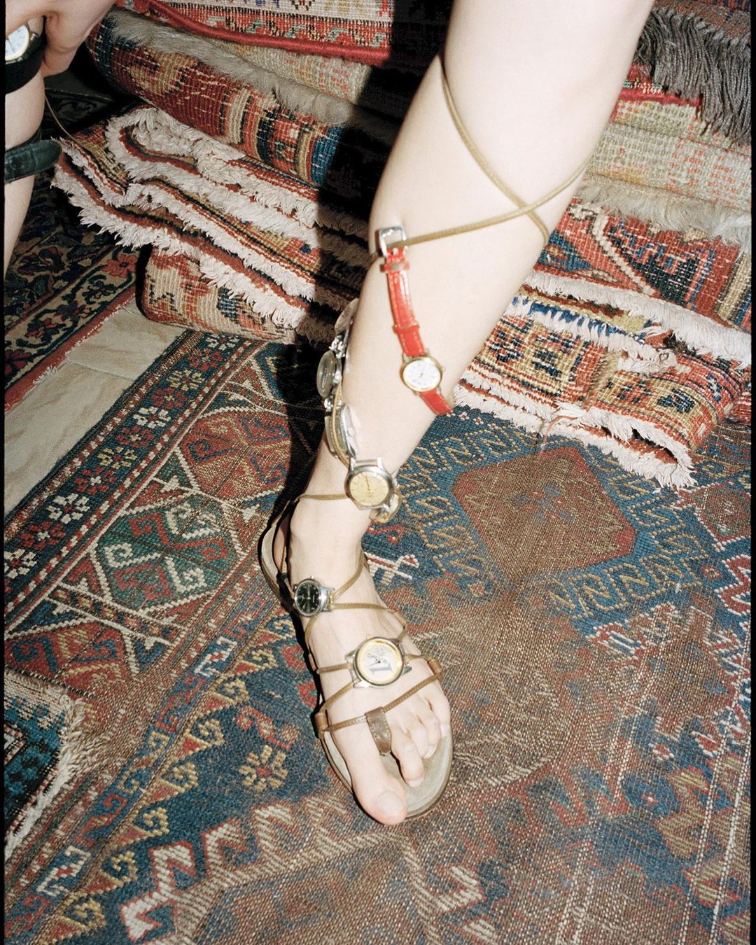Woman's leg adorned with multiple wristwatches worn as sandal accessories on a patterned rug, creative fashion statement.