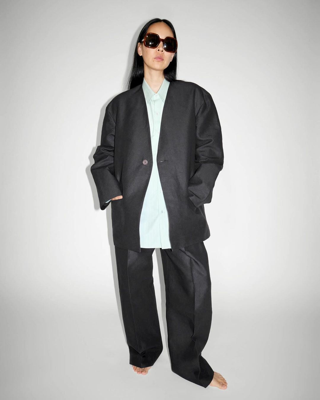 Adult woman poses in an oversized black suit and sunglasses against a bright neutral background in a fashion editorial-style portrait.