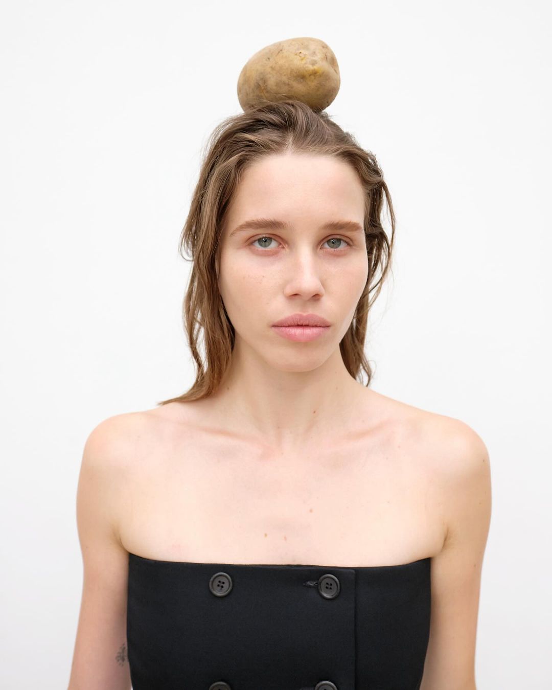 Portrait of a young woman wearing a black top with a potato balanced on her head against a white background in a studio shot.