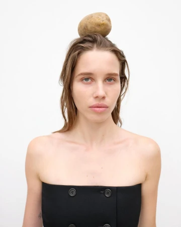 Portrait of a young woman wearing a black top with a potato balanced on her head against a white background in a studio shot.