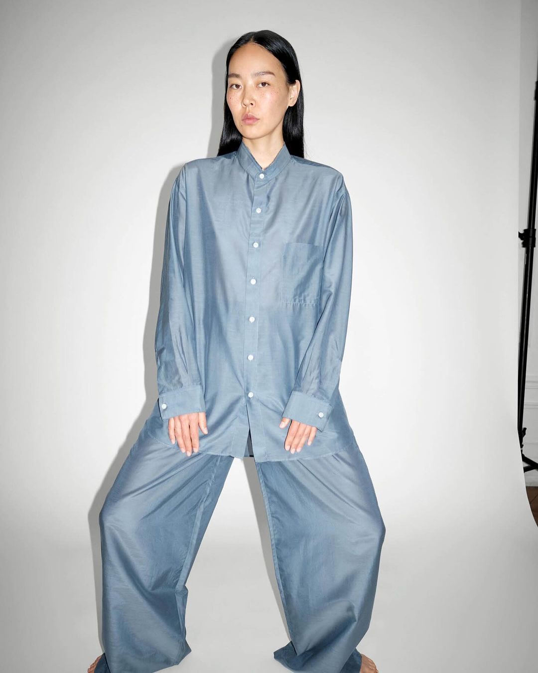 Asian woman in blue oversized shirt and pants in photo studio. She is standing with legs apart looking directly at the camera.