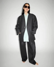 Model wearing large dark sunglasses, dark oversized blazer, trousers, and light shirt against white seamless background