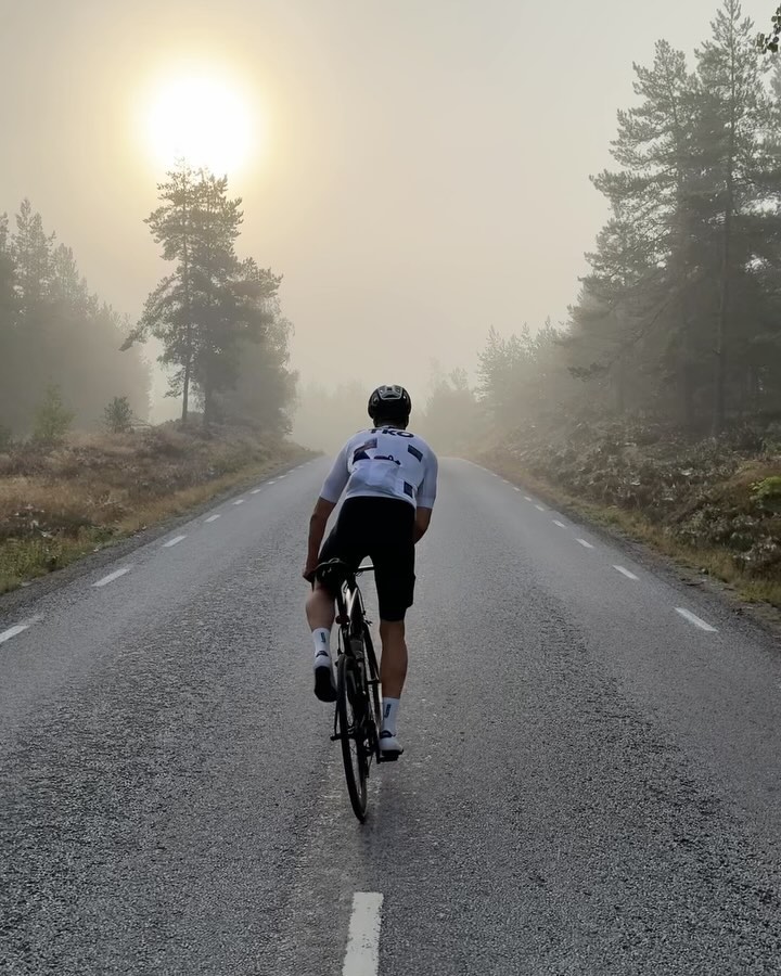 An adult male cyclist rides a bicycle on an asphalt road in a forest with fog and sunlight on a foggy morning.