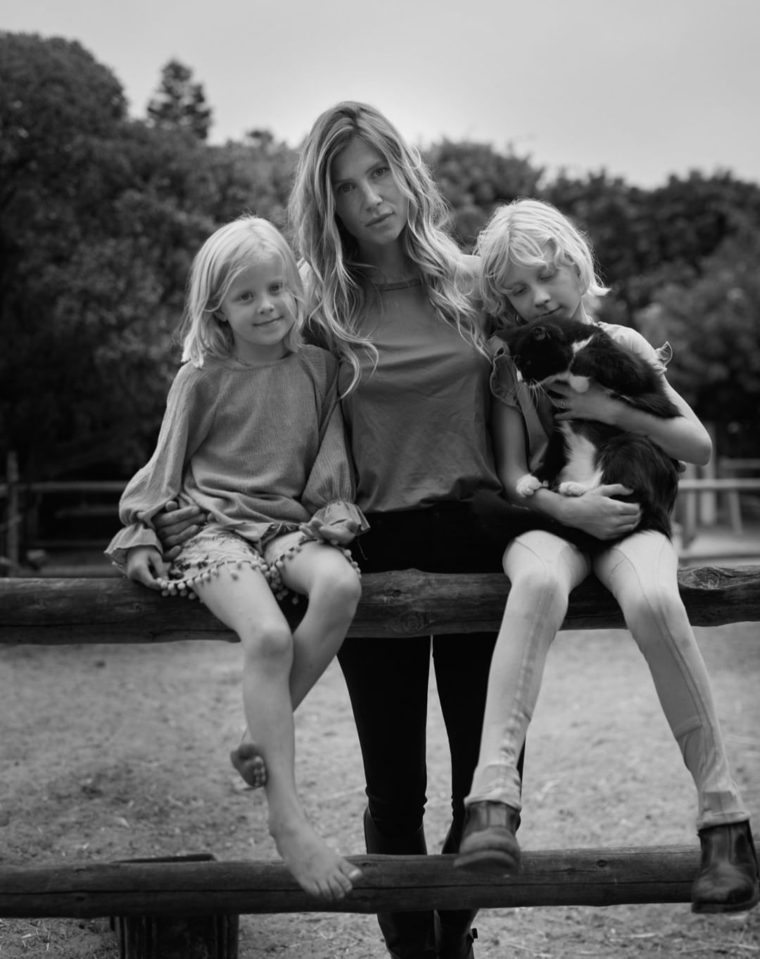 Black and white portrait of a mother and two daughters sitting on a wooden fence, with a dog, in an outdoor setting. 