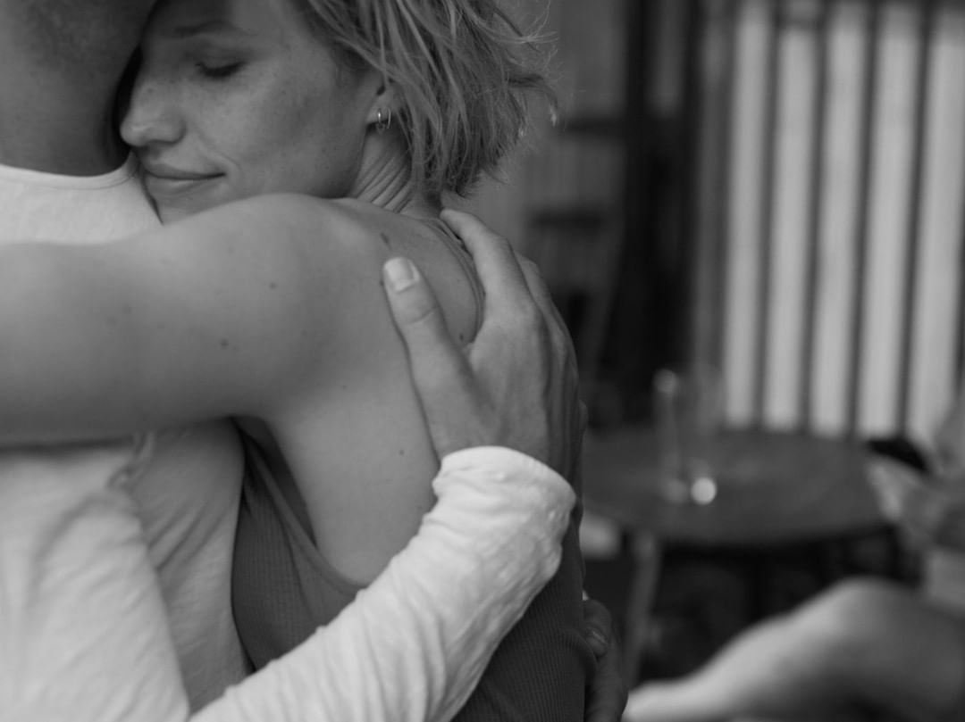 Close-up grayscale image of a woman being embraced by a man. The woman has short blonde hair and a tank top.