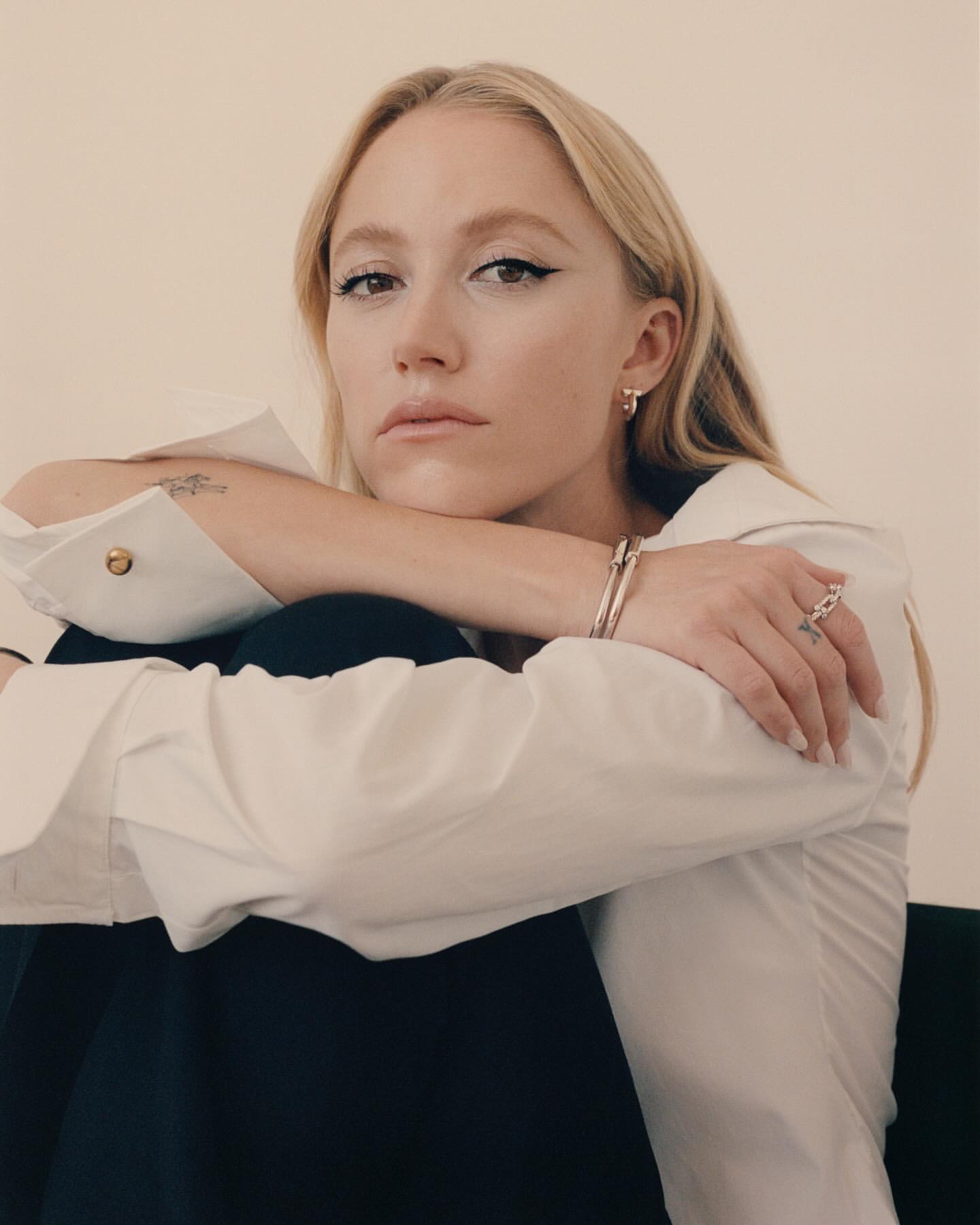 Blonde woman with arms crossed, wearing a white button-down shirt and black pants, looking directly at the camera in an indoor setting.