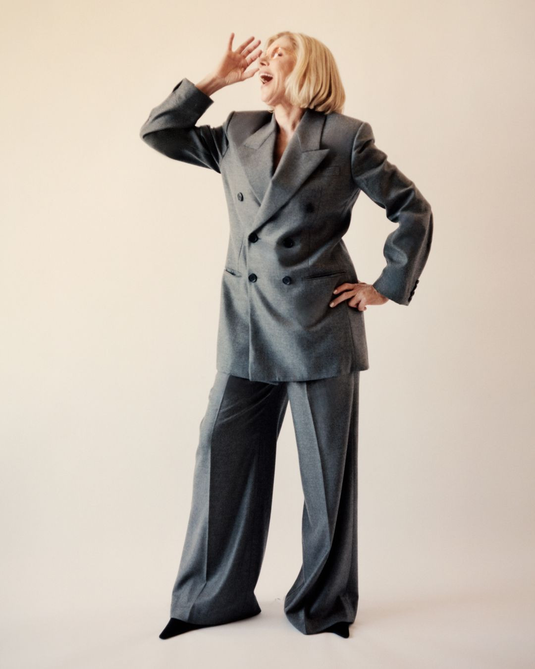 Full-shot of an older blonde woman in a gray double-breasted suit, standing with one hand on her hip and the other near her mouth, calling.