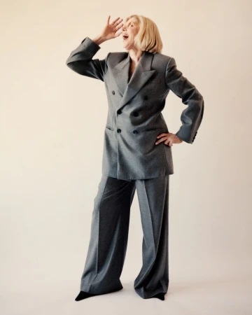Full-shot of an older blonde woman in a gray double-breasted suit, standing with one hand on her hip and the other near her mouth, calling.