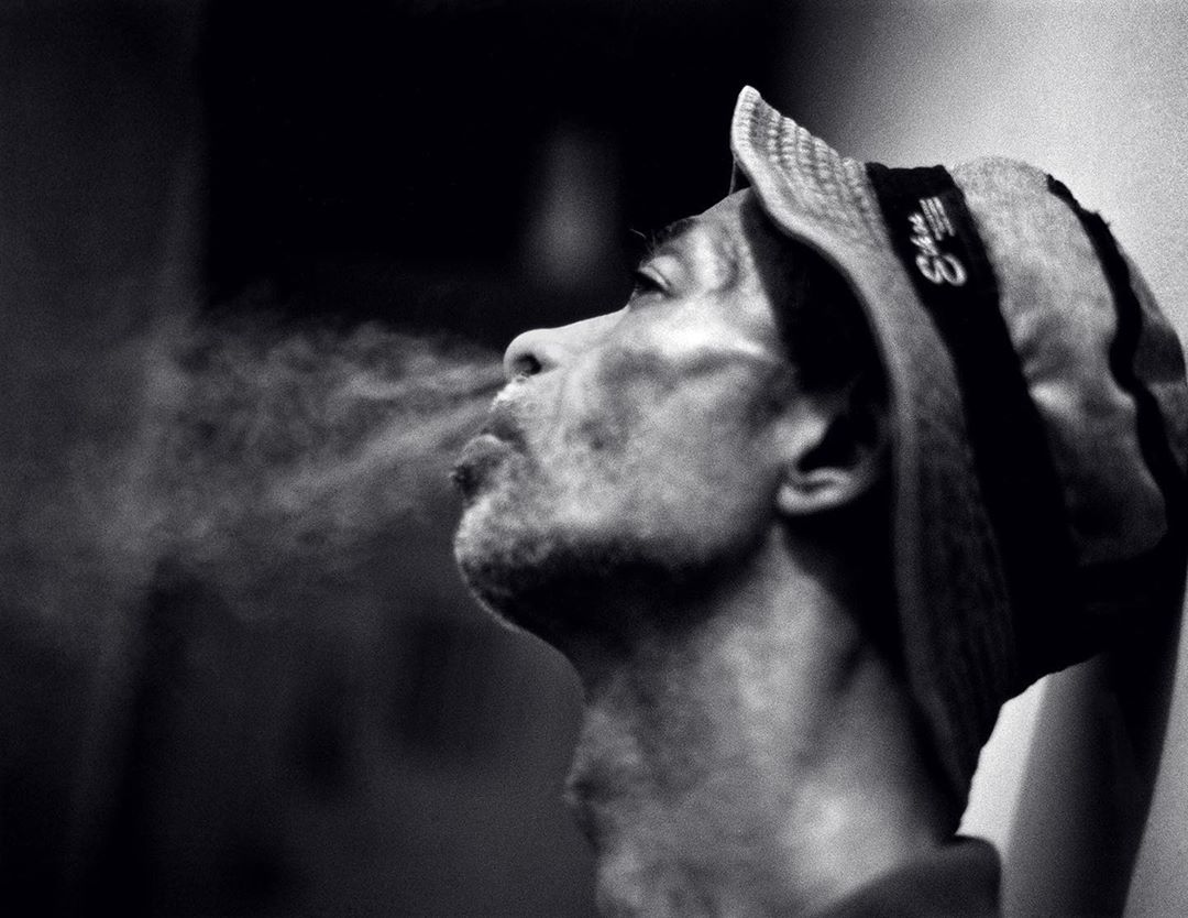 Monochromatic portrait of a person exhaling vapor into the air wearing a striped hat indoors in front of a dark background.