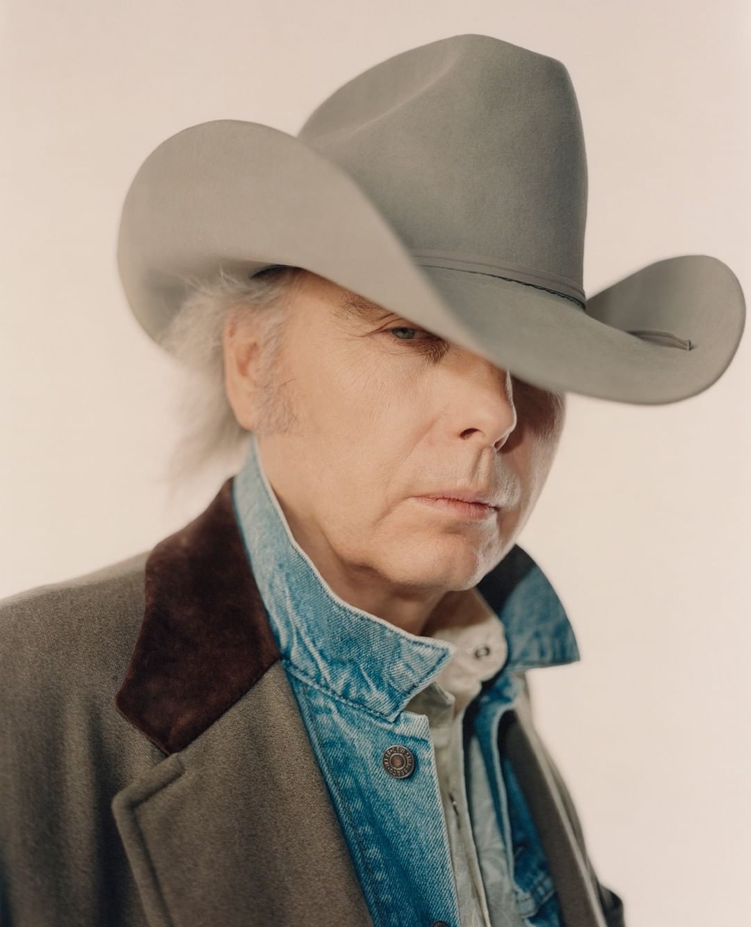 Portrait of a mature adult male wearing a grey cowboy hat and denim jacket, posing in a photo studio with a neutral background.