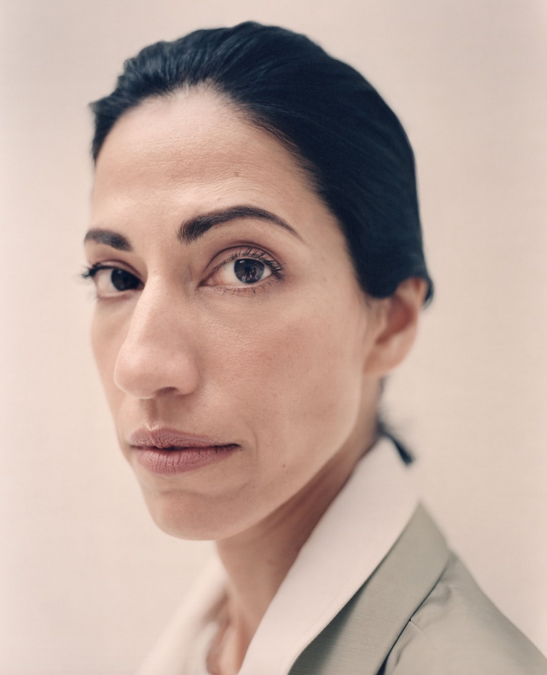 Close-up studio portrait of a woman with dark hair and olive skin wearing a light green jacket, facing the camera in a neutral expression.