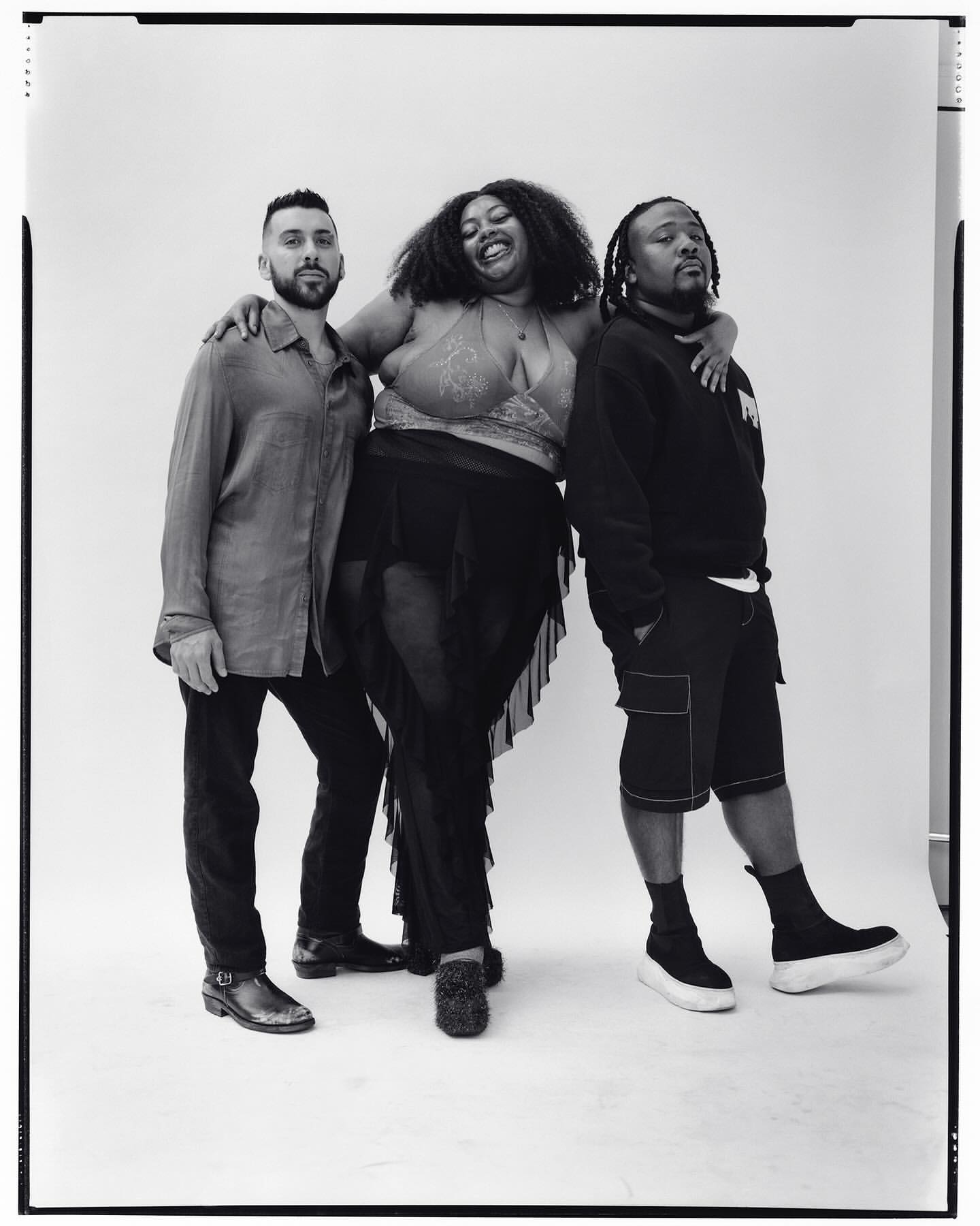 Three individuals pose together in a black and white studio shot. Two men flank a woman wearing a sparkly top and sheer black skirt, smiling.