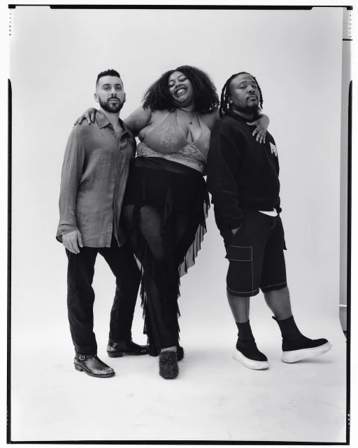 Three individuals pose together in a black and white studio shot. Two men flank a woman wearing a sparkly top and sheer black skirt, smiling.