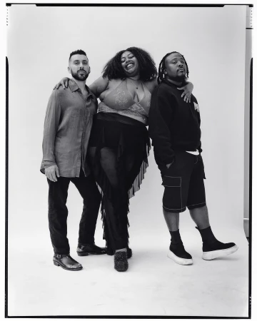 Three individuals pose together in a black and white studio shot. Two men flank a woman wearing a sparkly top and sheer black skirt, smiling.
