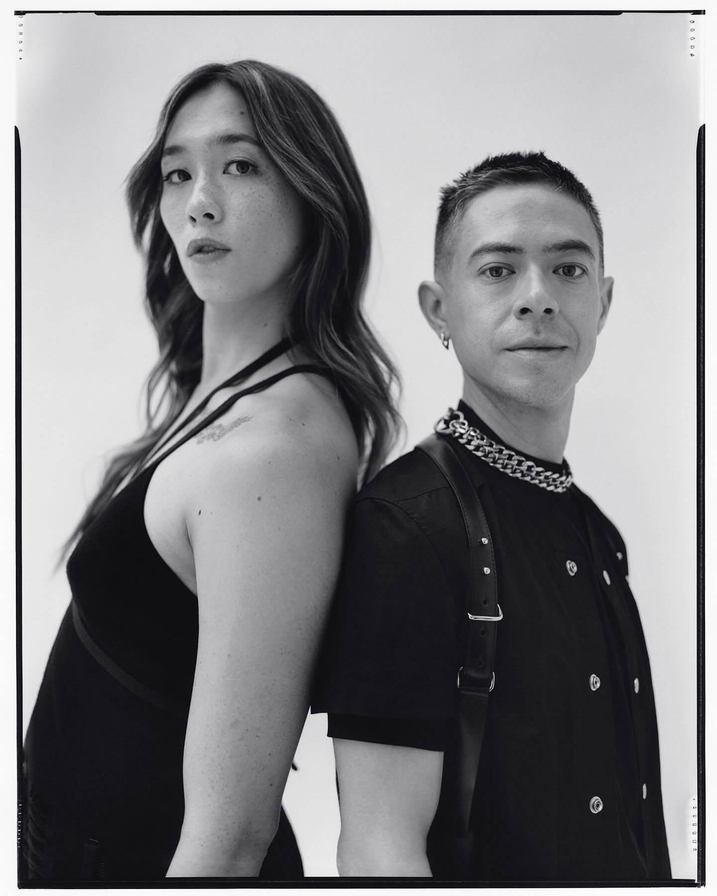 Monochrome portrait of two people standing back to back in a photo studio with a neutral background, creating a minimalist, fashion-focused image.