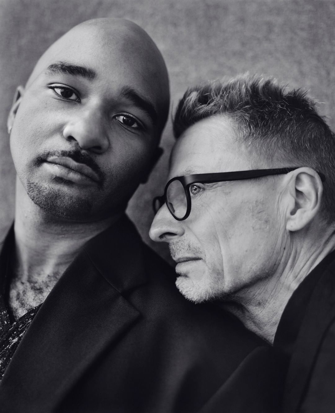 Portrait of two men in black jackets, one is bald and looking at the camera, the other is wearing glasses looking to the side.