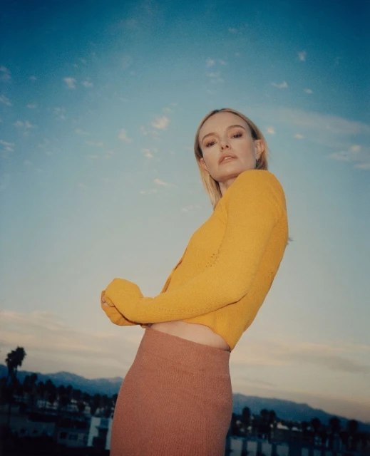 Woman in yellow top and skirt poses against a blue sky with palm trees and distant mountains during the daytime.