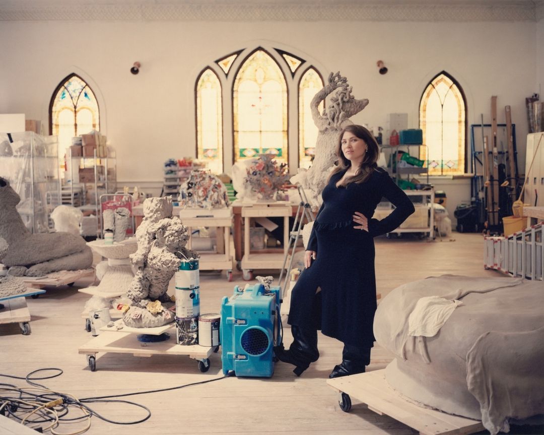 Pregnant woman in black dress poses inside a bright artist's studio surrounded by sculptures and art supplies beneath arched windows.