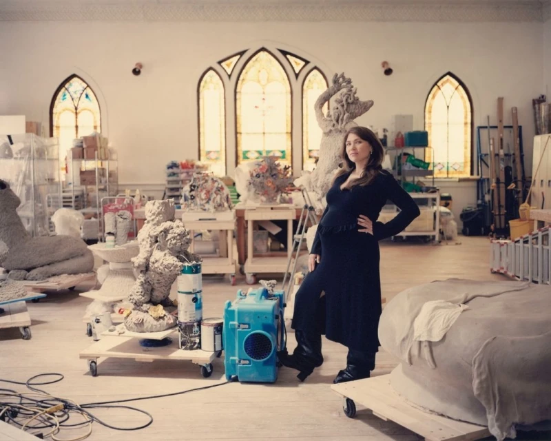 Pregnant woman in black dress poses inside a bright artist's studio surrounded by sculptures and art supplies beneath arched windows.