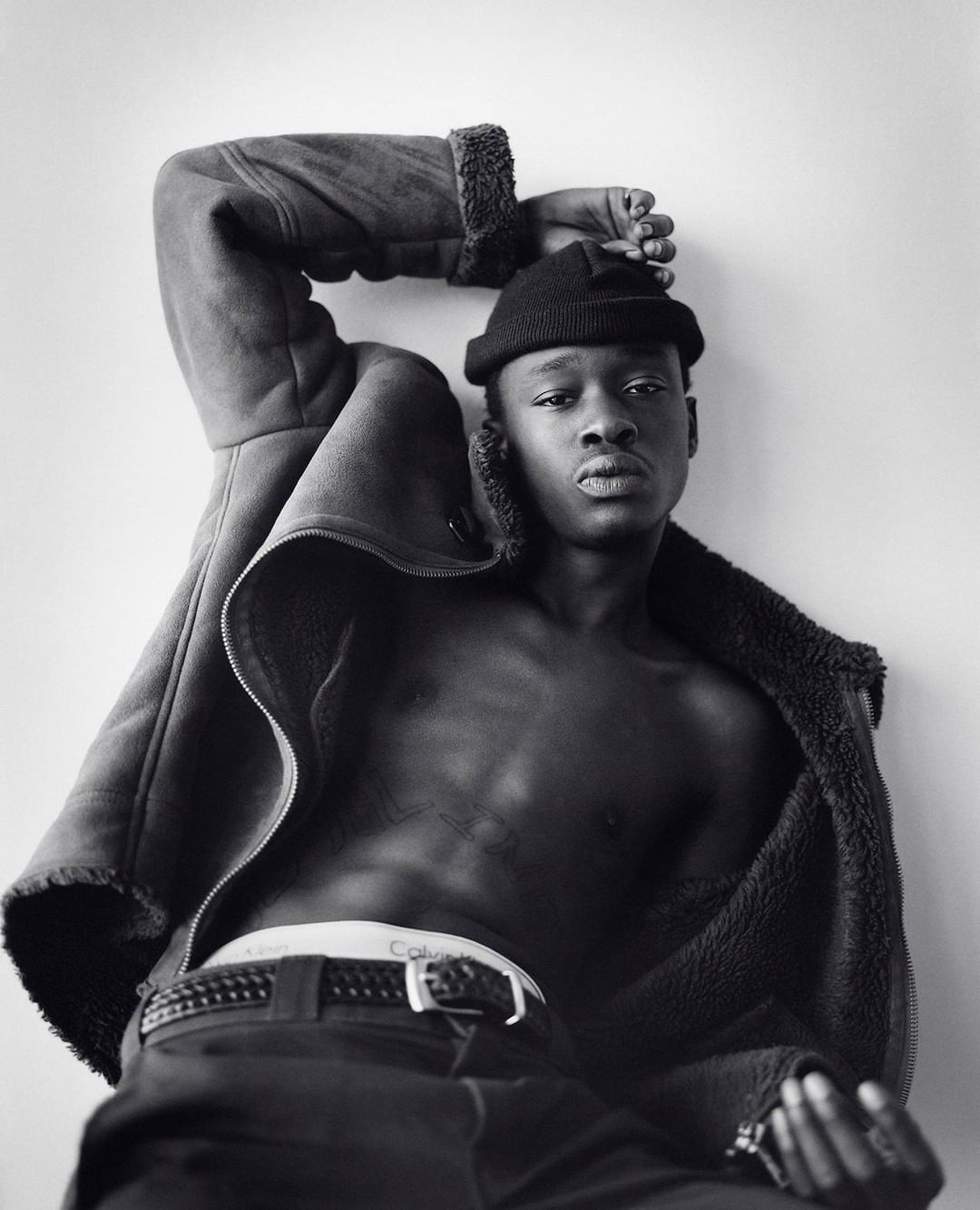 Monochrome portrait of a slender adult male with dark skin, wearing a black beanie and a coat, posing against a plain wall.
